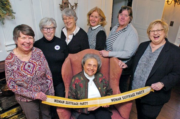 Members of the League of Women Voters 100th Anniversary Committee are, standing, left to right, Glorian Berk, Judy Samuelson, Ann Sandford, Cathy Peacock, Barbara McClancy, Susan Wilson and seated, Arlene Hinkemeyer at the Home Sweet Home Museum in East Hampton with an early 20th century suffrage sash. Missing from photo are committee members Judi Roth and
