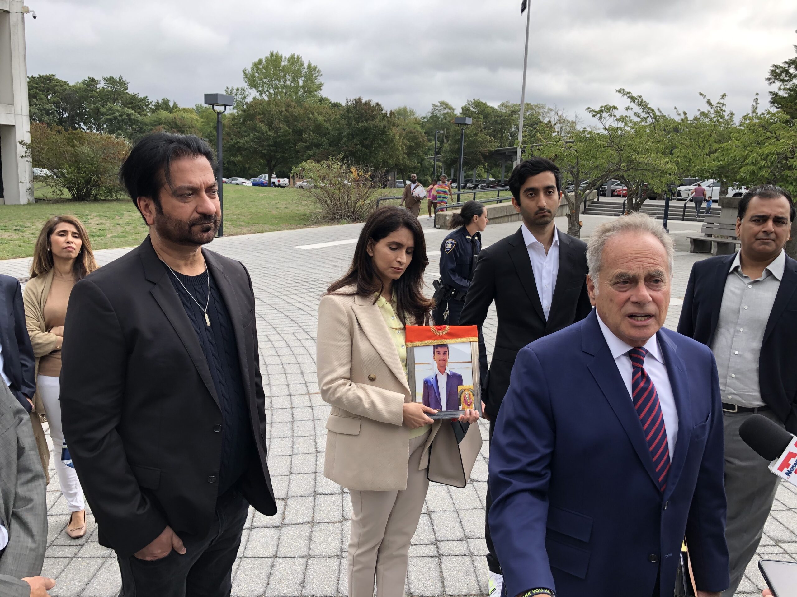 Benjamin Brafman, in the navy blue suit, speaks with the media outside the courthouse about the death of Devesh Kishore Samtani. The victim's mother, Mala Samtani, wearing a tan suit, stands behind Brafman, holding a photograph of her late son. To her right is the victim's father, Kishore Samtani. T.E. McMorrow