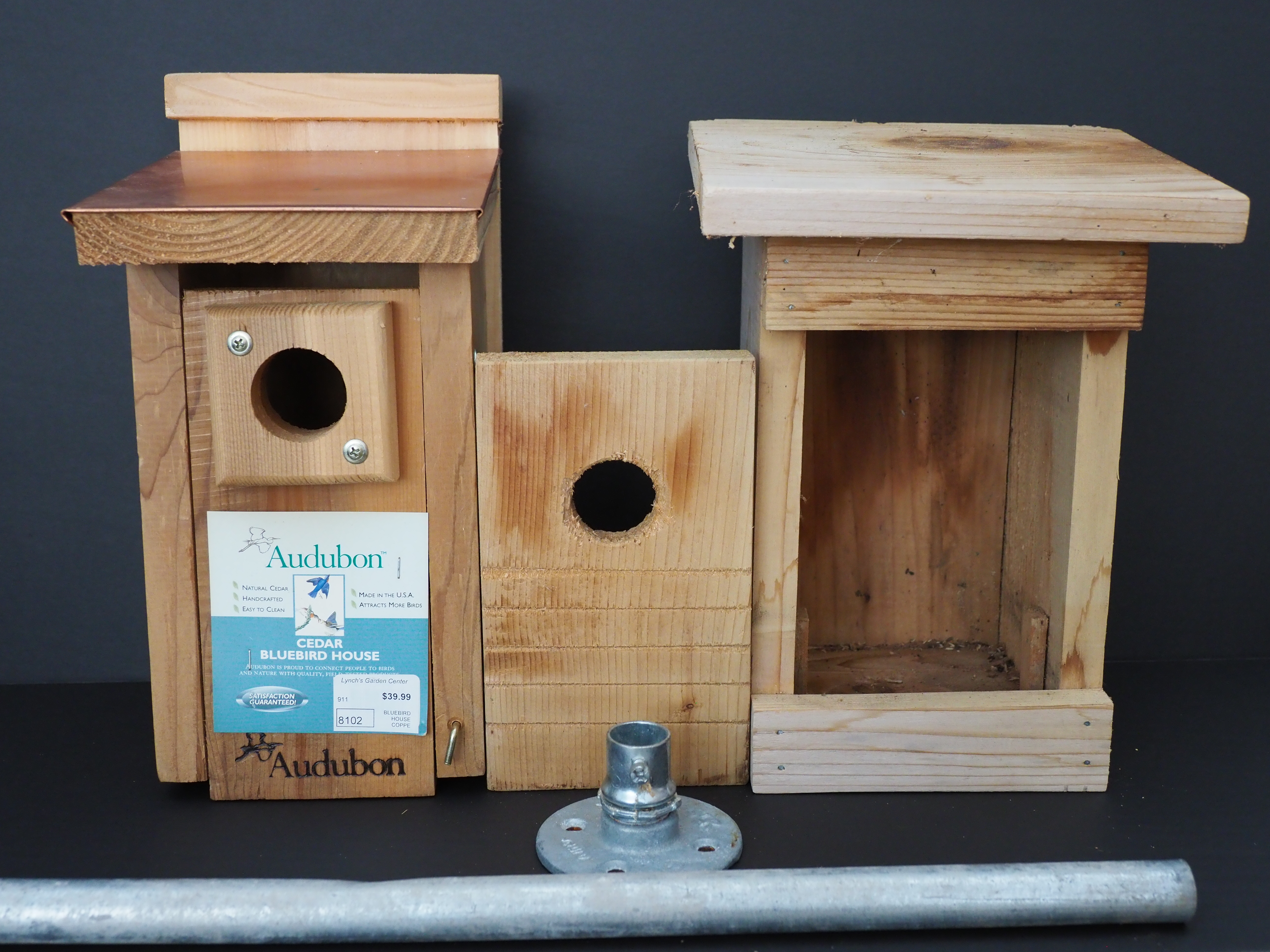 An Audubon-branded bluebird nesting box on the left and a homemade box on the right. The wood section in the center (reversed) shows the kerf cuts that allow the fledgling chick to climb up the wood when ready to leave the nest. The Audubon box does not have the kerf cuts and the copper covering on the top makes it too slippery for the birds to perch on top. The flange (bottom center) is attached with small bolts to the bottom of the box and the protruding connector allows easy mounting onto the EMT pipe. A simple set screw in the flange allows for easy removal of the entire box for repairs and cleaning.  The Audubon box is much heavier than the home made box and may not be supported by the EMT pipe. ANDREW MESSINGER